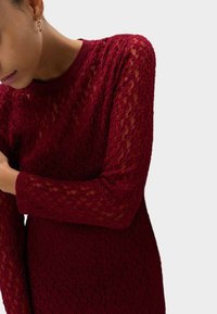 Rotes Spitzenkleid mit langen Ärmeln, rundem Halsausschnitt und strukturierter Oberfläche, das eine figurbetonte Silhouette und leicht durchscheinenden Stoff aufweist.