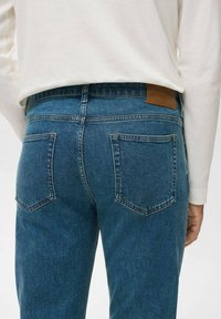 Jeans de mezclilla azul con un ajuste de cintura media, que cuentan con dos bolsillos traseros y una etiqueta de cuero marrón en la cintura. La textura parece resistente y duradera.