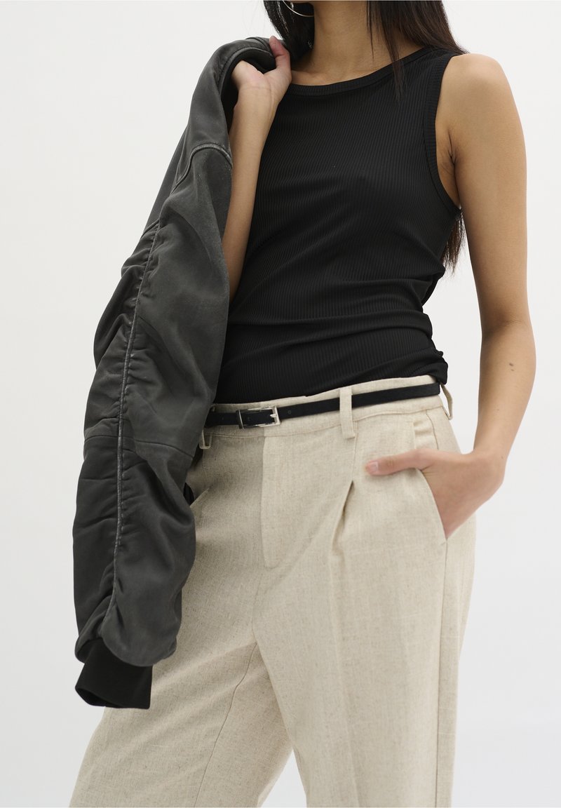 Femme portant un haut sans manches noir et un pantalon plissé beige, tenant une veste sombre sur l'épaule, main dans la poche du pantalon.