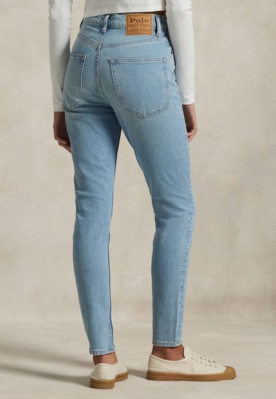 Persoon draagt lichtblauwe high-waist jeans en witte sneakers, gezien van achteren tegen een effen achtergrond.