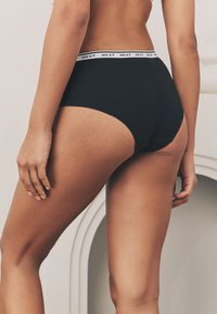Culottes en coton noir taille haute avec une ceinture blanche portant le logo "NEXT", présentant une texture lisse et un design couvrant entièrement.