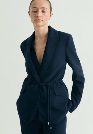 Mujer con blazer azul marino a medida con bolsillos delanteros y un cinturón fino, parada con las manos en los bolsillos frente a un fondo liso.