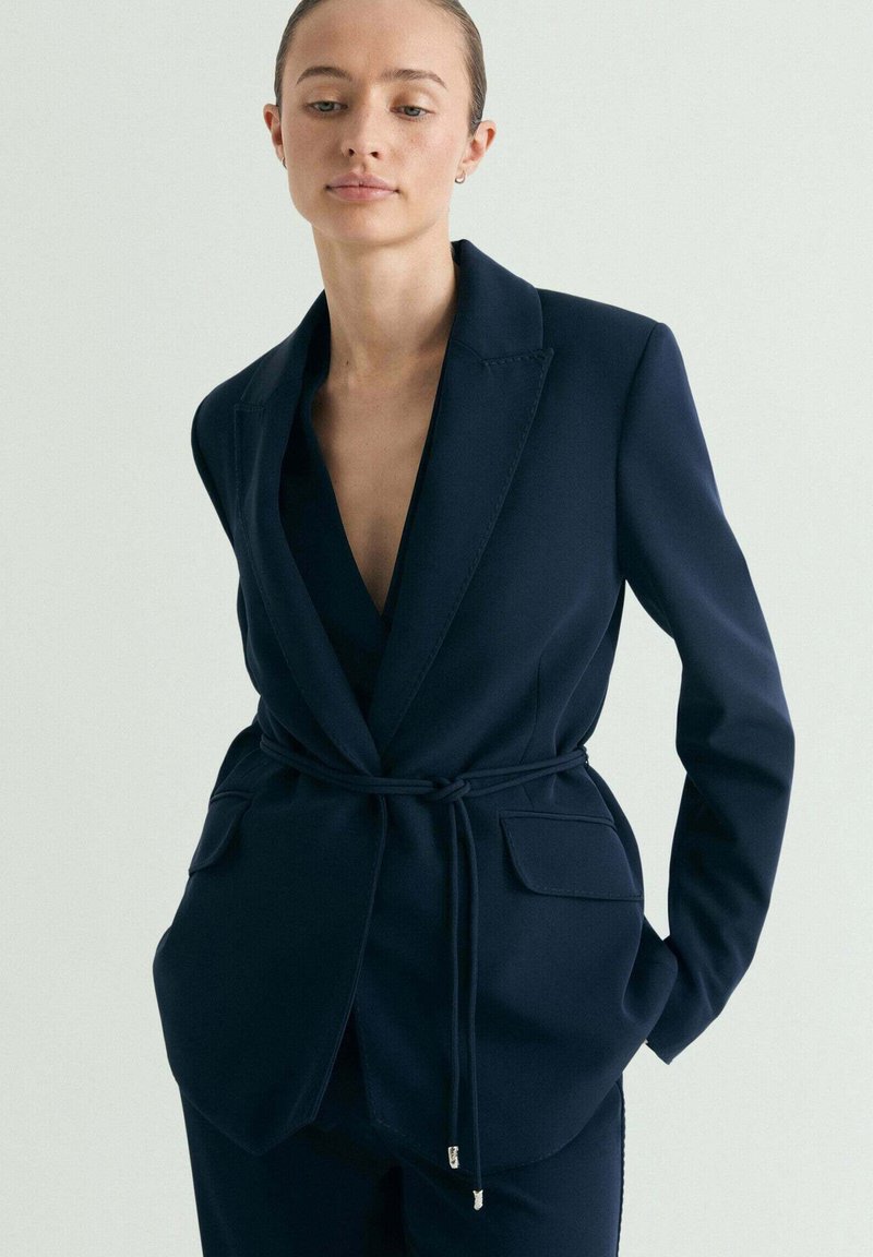 Woman wearing a tailored navy blazer with front pockets and a thin belt, standing with hands in pockets against a plain background.