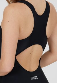 Maillot de bain noir avec un design dos nu et un dos ouvert. Texture lisse avec un léger éclat, rehaussé par un petit logo.