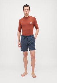 Brun kortermet rashguard med logo på brystet, kombinert med mørkeblå svømshorts med elastisk liv og snøring.