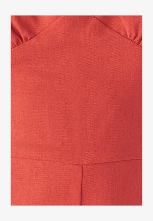 Close-up van koraalkleurige stof met zichtbare weefstructuur en naaddetails op een kledingstuk met geraffineerde bovenranden.