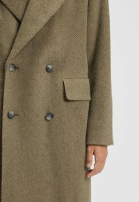 Double-breasted olive green coat made of herringbone fabric. Features dark buttons and a chest pocket. Textured, warm, with a tailored fit.