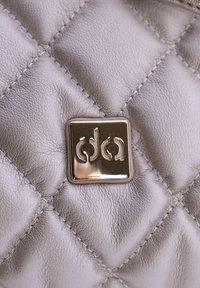 Cuero gris acolchado con un emblema rectangular metálico. Presenta un patrón de costura y una textura suave, enfatizando la profundidad y el detalle.