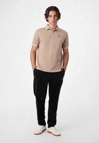 Polo beige con cuello a contraste y pequeño logo en el pecho, combinado con pantalones negros y zapatos blancos sobre un fondo liso.