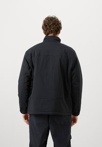 Man with curly hair wearing a black padded jacket and black pants, standing facing away against a plain light background.