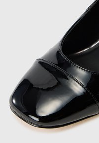 Detailný záber čiernej lakovanej topánky so štvorcovou špičkou a precíznym prešívaním na bielom pozadí.