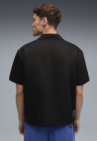 Sort kortærmet skjorte med flad krave, glat tekstur og mærkeetiket på siden, båret over lyseblå shorts.