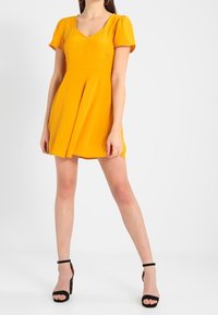 Gul klänning med korta puffärmar, V-ringning och utsvängd kjol. Bärs med svarta högklackade sandaler med ankelband.