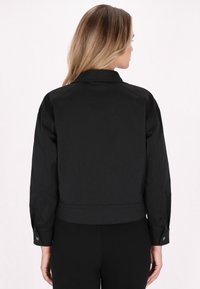 Jachetă scurtă neagră cu guler, având mâneci lungi și manșete cu butoane. Textură netedă și design simplu, fără modele sau embellishment-uri vizibile.
