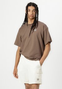 Brūns īss piedurkņu T-krekls ar baltu Adidas logo, kombinēts ar gaiši beidžiem šortiem ar sānu kabatu un elastīgu jostasvietu.