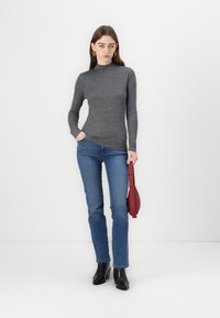 Grå langermet topp med høy hals, kombinert med blå jeans. Modellen holder en rød håndveske og har på seg svarte ankelstøvler. Minimalistisk bakgrunn.
