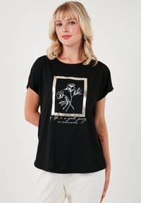 Zwarte t-shirt met een losse pasvorm, voorzien van een wit bloemenpatroon binnen een gouden rechthoekig kader en tekst eronder. Zachte stof textuur.