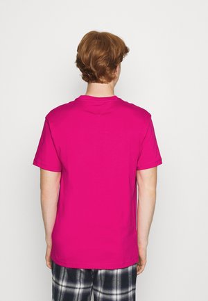 Person with short brown hair facing away, wearing a bright pink short-sleeve shirt and black-and-white plaid pants against a plain background.