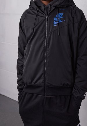 Personne portant une veste noire zippée Nike avec un logo bleu sur la poitrine et un pantalon noir, debout les mains dans les poches contre un mur gris.