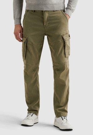 Muž v olivových cargo nohaviciach s viacerými vreckami a bielych teniskách, ruky vreckách, na jednofarebnom pozadí.