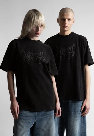 Δύο άτομα που φορούν μαύρα γραφικά μπλουζάκια και μπλε τζιν στέκονται μπροστά σε ένα απλό ανοιχτό φόντο με ουδέτερες εκφράσεις.