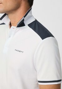 Bílá polokošile s námořnickými modrými akcenty, zvýšeným límcem a kontrastními pruhy. Na hrudi je vyšívané logo 'HACKETT'.