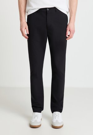 Homme portant un pantalon noir ajusté, des baskets blanches et une chemise blanche, les mains détendues le long du corps sur un fond uni.