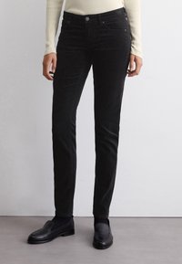 Pantalon en velours côtelé noir à coupe slim et texture subtile, avec une fermeture à bouton sur le devant et des poches latérales, associé à des mocassins noirs.