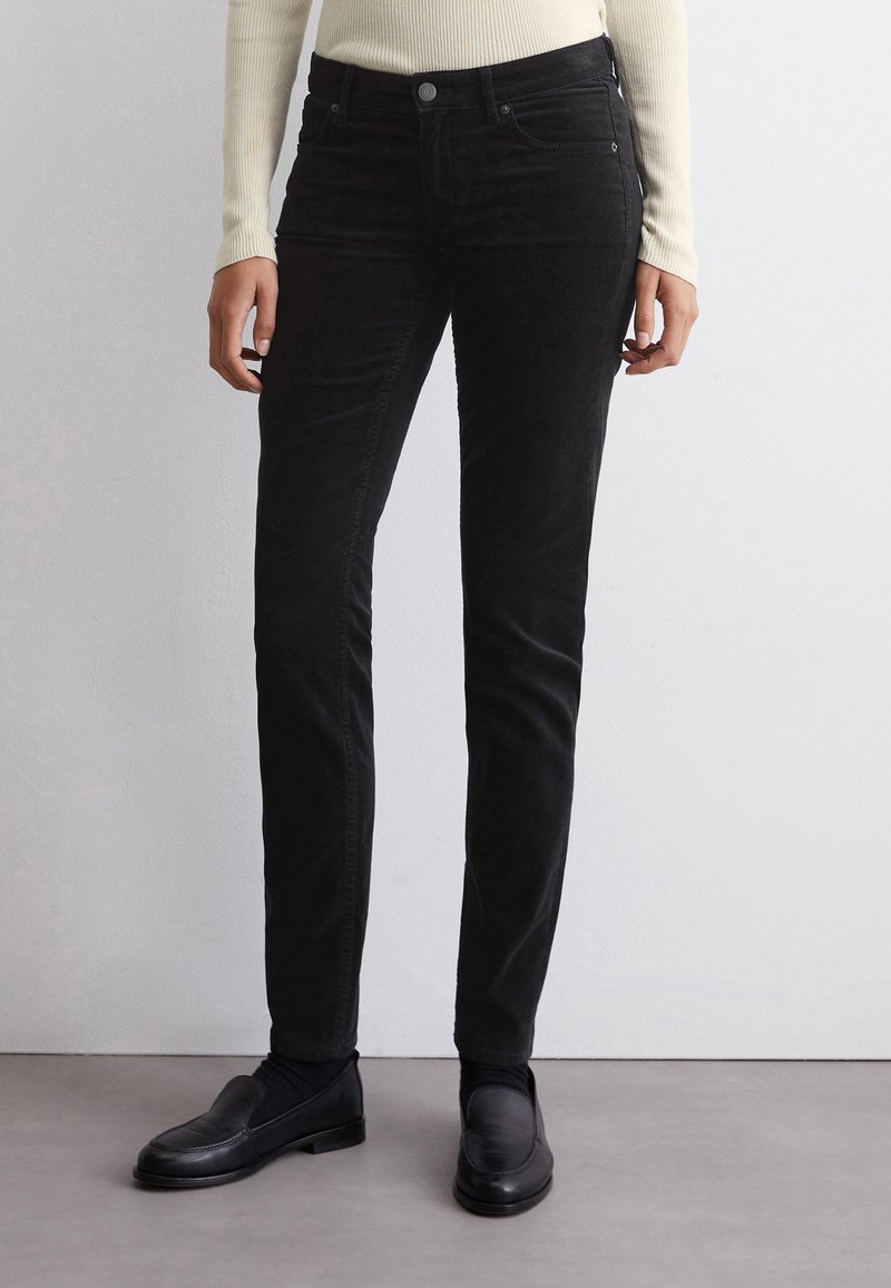 Pantalon en velours côtelé noir à coupe slim et texture subtile, avec une fermeture à bouton sur le devant et des poches latérales, associé à des mocassins noirs.