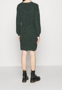 Stickad klänning i mörkgrön, figurnära med lång puffärmar och rund halsringning. Bärs med svarta kängor som har gula detaljer.