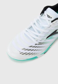 Zapatilla deportiva blanca con acentos en negro y verde azulado; cuenta con un diseño texturizado, material sintético en capas y una suela con patrón.