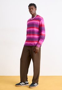 Överdimensionerad, ribbad stickad tröja med rosa och lila horisontella ränder, kombinerad med bruna mönstrade byxor och svarta sneakers med vita ränder.