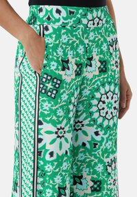 Grüne weit geschnittene Hose mit einem Blumen- und geometrischen Muster, glattem Stoff, elastischem Bund und seitlichen Taschen mit kontrastierender Paspel.