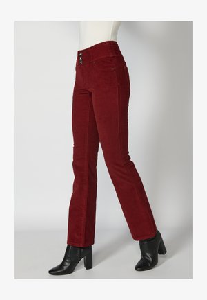 Pantalon évasé en velours côtelé rouge avec une taille haute, doté de trois boutons décoratifs et de poches avant, associé à des bottines à talons noires.