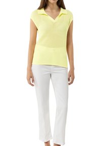 Żółta siateczkowa koszulka polo z dekoltem w serek, krótki rękaw typu cap, noszona z dopasowanymi białymi spodniami. Widoczne detale teksturowanej tkaniny i kołnierzyka.