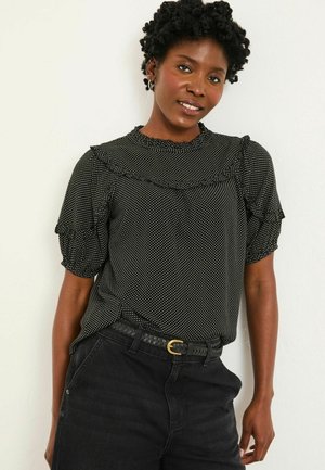 Schwarz-weiße Bluse mit Pünktchenmuster, Puffärmeln, rüschigem Ausschnitt und weicher Textur. Kombiniert mit hochgeschnittenen schwarzen Hosen und einem geflochtenen Gürtel.