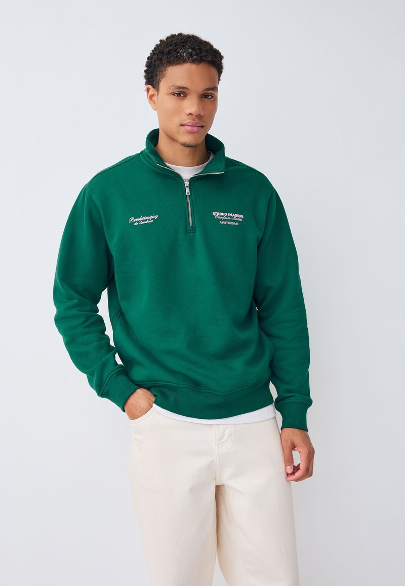 Grøn sweatshirts med lynlås lavet af blødt stof, med lange ærmer, høj krave og broderet tekst foran. Kombineret med beige bukser.