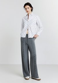 Chemise blanche à boutons avec des rayures grises, associée à un pantalon gris ample avec une ceinture élastique et des poches latérales. Chaussures de couleur claire.