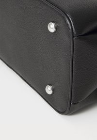 Esquina inferior de una bolsa de cuero negro texturizado con dos remaches de metal plateado sobre una superficie blanca.