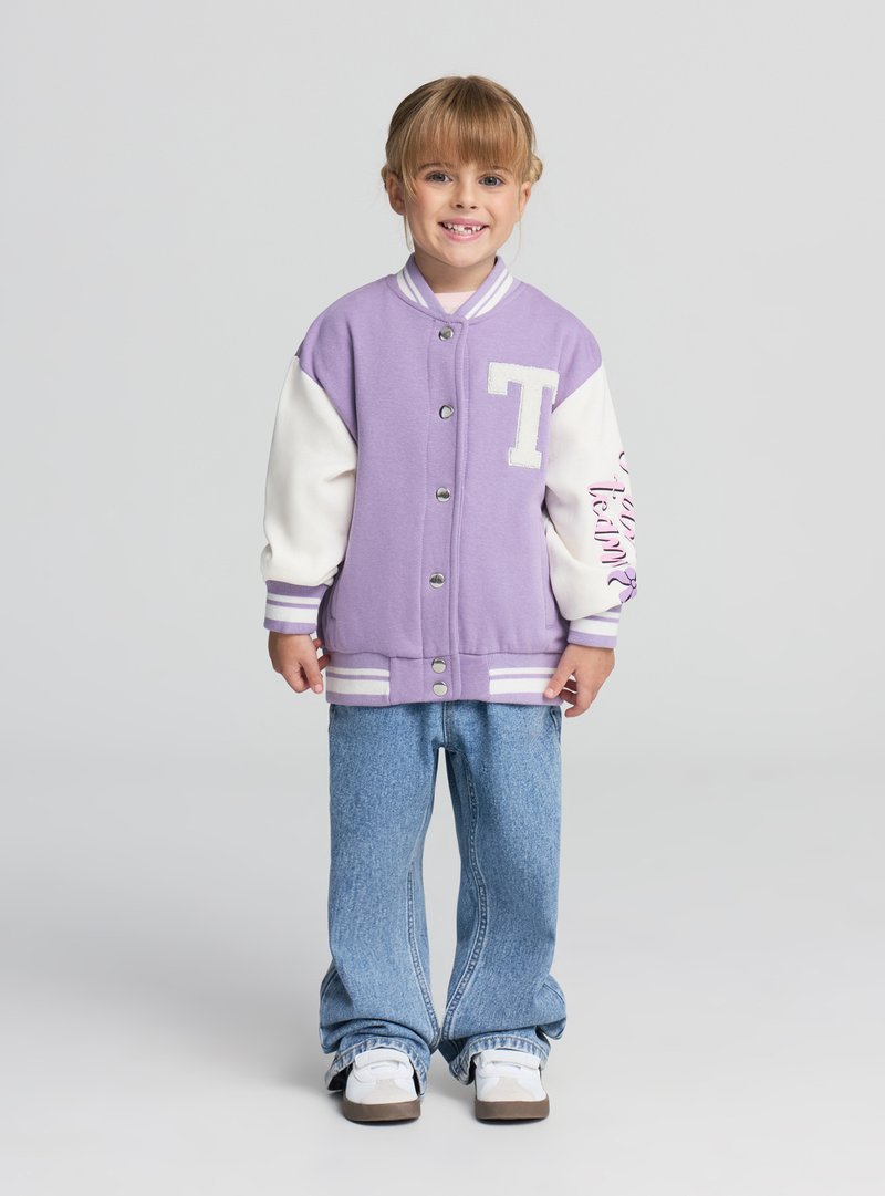 Lawendowa kurtka typu varsity z białymi rękawami, zapinana na guziki z przodu, z logo; zestawiona z jasnoniebieskimi szerokimi spodniami i białymi trampkami.