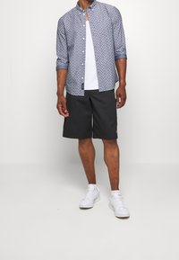 Košile s krátkým rukávem, zapínaná na knoflíky, s modrým geometrickým vzorem, v kombinaci s černými šortkami po kolena a bílými teniskami.