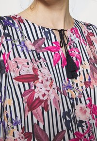 Bluzka w kwiatowy wzór z pionowymi czarnymi i białymi paskami, z wiązaniem przy dekolcie i czarnym frędzlami. Miękka tekstura materiału.