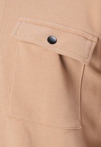 Tasca in tessuto beige con pattina fissata da un singolo bottone a pressione nero, che mostra una trama testurizzata e dettagli di cucitura.