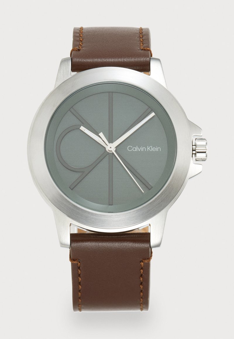 Montre en acier inoxydable argenté avec un cadran vert, un bracelet en cuir marron et des accents argentés. Elle présente un design minimaliste avec un logo et des index structurés.
