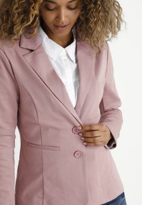 Blazer rose avec une finition texturée, design à un bouton, et deux boutons. Accompagné d'une chemise blanche à col en dessous.
