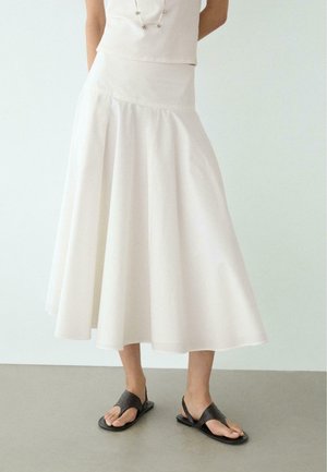 Frau trägt einen weißen Midirock, ein weißes Oberteil, eine Halskette und schwarze flache Sandalen, steht auf einem glatten grauen Boden vor einem schlichten Hintergrund.