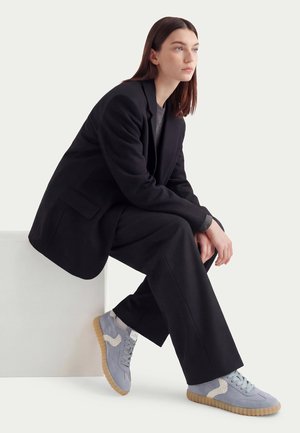 Young woman with long brown hair sits on white block, wearing black oversized suit and light blue sneakers with gum soles.