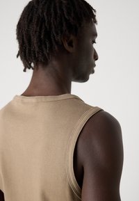 Top bej fără mâneci, cu o textură în dungi, având un guler rotund și o croială lejeră, evidențiată de tonul uniform al pielii modelului.