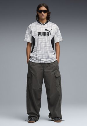 Persoon die een wit Puma sportshirt, donkere oversized cargobroek, zwarte sneakers en zwarte zonnebril draagt, staand tegen een grijze achtergrond.