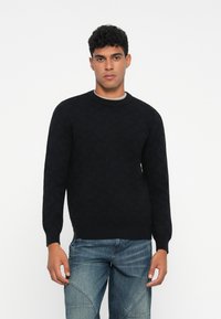 Mann in einem schwarzen gemusterten Langarmpullover und blauen Jeans, der vor einem weißen Hintergrund steht.